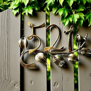 Op de afbeelding: Decoratieve metalen huisnummers 521 in goud, gemonteerd op een grijze houten hek. De cijfers zijn gestileerd met een bloemenrank en bladeren, met groene bladeren op de achtergrond.