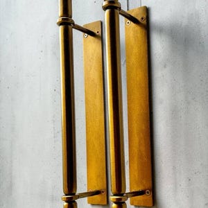 K&ouml;nnte beinhalten: Zwei vertikale T&uuml;rgriffe aus Messing mit zylindrischem Stangendesign, abgerundeten Endst&uuml;cken oben und unten sowie rechteckigen Montageplatten. Jeder Griff ist mit zwei horizontalen Halterungen und sichtbaren Schrauben an der Platte befestigt.