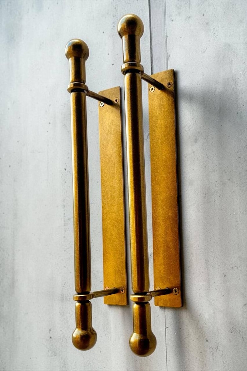 K&ouml;nnte beinhalten: Zwei vertikale T&uuml;rgriffe aus Messing mit zylindrischem Stangendesign, abgerundeten Endst&uuml;cken oben und unten sowie rechteckigen Montageplatten. Jeder Griff ist mit zwei horizontalen Halterungen und sichtbaren Schrauben an der Platte befestigt.