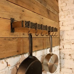 Puede incluir: Un estante rústico para ollas de cocina con siete ganchos, montado en una pared de madera. Dos ollas cuelgan de los ganchos: una sartén de hierro fundido negro y una cacerola de acero inoxidable. La pared es de madera reciclada y ladrillo blanco.