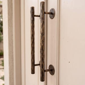 May include: A vertical door handle with a dark bronze, hammered metal texture. The handle is mounted to a light-colored wooden door with two circular plates and screws. Below the handle, a small metal door knocker and a keyhole plate are visible.