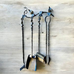 May include: A black wrought iron fireplace tool set with a decorative top featuring blue and gold accents. The set includes a poker, shovel, brush, and a tool with a looped handle. The tools hang on a matching holder.