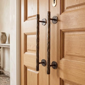 May include: A pair of vertical door handles with a dark, hammered metal texture, mounted on a light-colored wooden door. The handles feature circular mounting plates secured with screws, and a brass-colored keyhole plate is positioned above them.