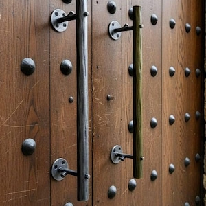 Puede incluir: Primer plano de una puerta de madera con dos manijas metálicas. La puerta está hecha de tablones de madera verticales y está tachonada con numerosos acentos metálicos oscuros y redondeados. Las manijas son cilíndricas, una gris oscuro y la otra verde oliva.