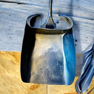 May include: A hand-forged metal dustpan with a long handle, showcasing a curved design and a textured surface. The dustpan is made of dark metal and is hanging on a wooden surface.