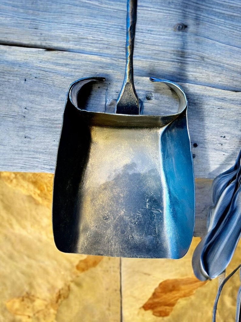 May include: A hand-forged metal dustpan with a long handle, showcasing a curved design and a textured surface. The dustpan is made of dark metal and is hanging on a wooden surface.