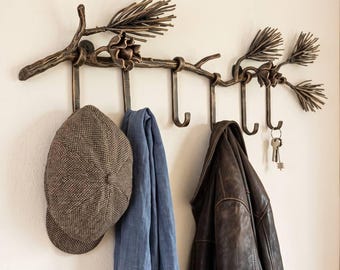 May include: A wall-mounted coat rack shaped like a bronze-toned pine branch with needles and cones. It features six hooks holding a grey herringbone cap, a blue scarf, a brown leather jacket, and a set of silver keys hanging from the end hook.