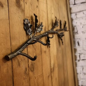 May include: A handcrafted metal wall hook shaped like a tree branch with leaves. The hook is mounted on a wooden surface. The metal has a dark, rustic finish, and the wood has a natural grain.