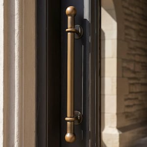 May include: A bronze door pull handle with a cylindrical design and rounded ends. The handle is mounted on a dark grey door, with a glimpse of a stone building in the background. The handle is a vertical bar with a circular ring in the middle.