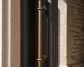 Könnte beinhalten: Ein bronzefarbener Türgriff mit zylindrischem Design und abgerundeten Enden. Der Griff ist an einer dunkelgrauen Tür montiert, mit einem Blick auf ein Steingebäude im Hintergrund. Der Griff ist ein vertikaler Stab mit einem kreisförmigen Ring in der Mitte.