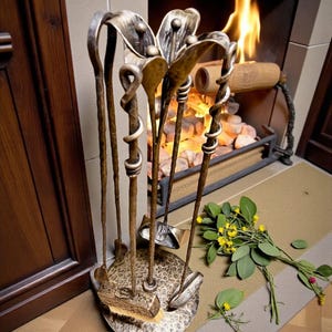 May include: A decorative fireplace tool set with a floral design. The set includes a poker, shovel, brush, and tongs, all made of metal with a bronze finish. The tools are arranged on a round base with a hammered texture. A bouquet of yellow flowers and greenery rests nearby.