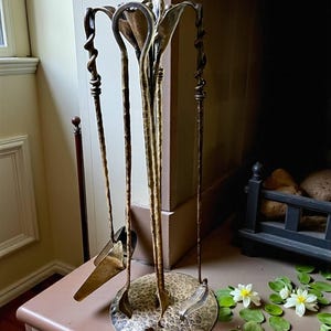 Pode incluir: Um conjunto de ferramentas decorativas para lareira com base em latão martelado e ferramentas a condizer. O conjunto inclui um atiçador, uma pá e um tenaz, todos com cabos ornamentados e torcidos. As ferramentas estão dispostas num suporte vertical.