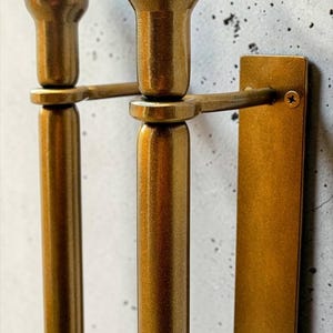 K&ouml;nnte beinhalten: Ein wandmontierter Handtuchhalter aus messingfarbenem Metall mit zwei vertikalen zylindrischen Stangen, die mit kugelf&ouml;rmigen Endst&uuml;cken versehen sind. Die Stangen sind &uuml;ber horizontale Halterungen an einer rechteckigen Platte befestigt.