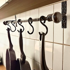 May include: A wrought iron kitchen pot rack with several S-hooks holding black frying pans. The rack is mounted on white tiled wall. The rack has a hammered texture and is attached to the wall with decorative brackets.