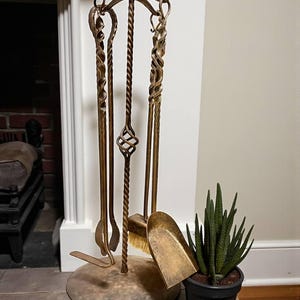 Peut inclure: Un ensemble d'outils de cheminée en laiton avec un support. L'ensemble comprend un tisonnier, une pelle, une brosse et des pinces. Les outils ont des poignées torsadées décoratives et sont disposés sur une base ronde. Une plante en pot est à côté de l'ensemble d'outils.