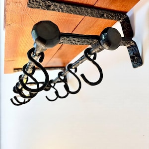 May include: A wall-mounted shelf with a natural wood finish and black metal detailing. The shelf has a series of hooks for hanging items. The design combines wood and metal for a vintage aesthetic, suitable for a kitchen or hallway.