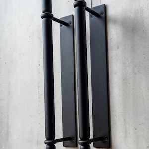 K&ouml;nnte beinhalten: Zwei matt-schwarze T&uuml;rgriffe aus Metall mit zylindrischem Stangendesign und kugelf&ouml;rmigen Endst&uuml;cken oben und unten. Jeder Griff ist auf einer rechteckigen Platte montiert, die mit sichtbaren Schrauben befestigt ist und ein klares Design aufweist.