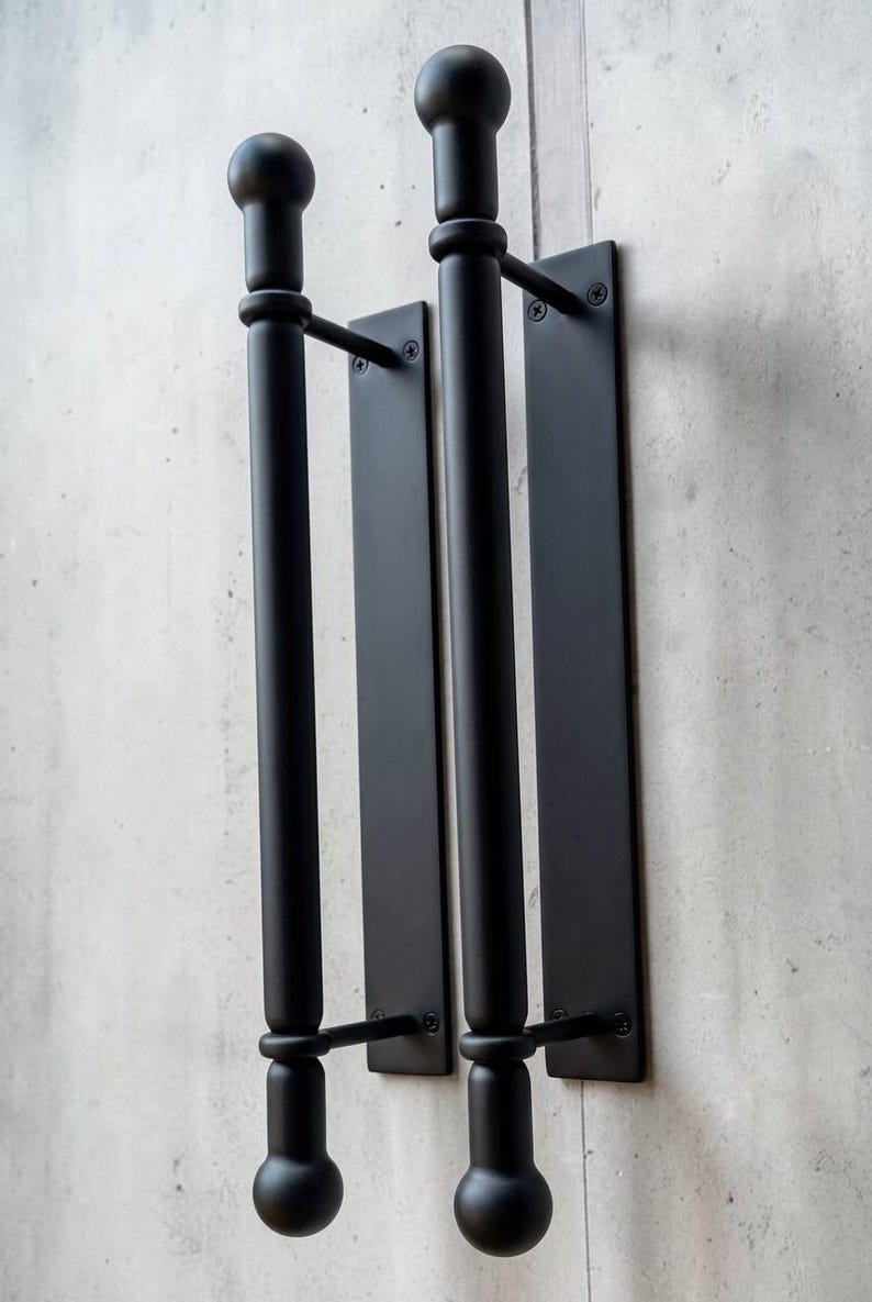 K&ouml;nnte beinhalten: Zwei matt-schwarze T&uuml;rgriffe aus Metall mit zylindrischem Stangendesign und kugelf&ouml;rmigen Endst&uuml;cken oben und unten. Jeder Griff ist auf einer rechteckigen Platte montiert, die mit sichtbaren Schrauben befestigt ist und ein klares Design aufweist.