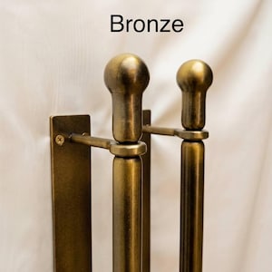 K&ouml;nnte beinhalten: Ein bronzefarbener Handtuchhalter aus Metall zur Wandmontage mit zwei vertikalen Stangen und abgerundeten Endst&uuml;cken. Die Halterung besteht aus einer rechteckigen Montageplatte mit einer sichtbaren Schraube. Das Wort Bronze steht dar&uuml;ber.