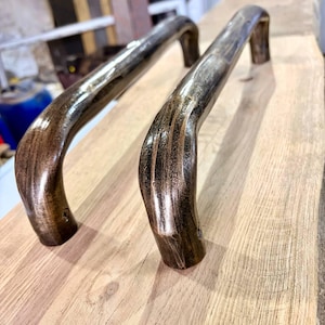 May include: Two bronze-colored metal door handles with a curved design. The handles are placed on a light-colored wooden surface. The handles have a rustic, aged appearance, suggesting they are handcrafted.