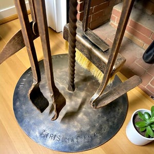 May include: A wrought iron fireplace tool set with a circular base. The set includes a poker, shovel, brush, and tongs. The base is inscribed with "CHRISTMAS 2020". The tools have a dark, aged finish, and the brush has yellow bristles.