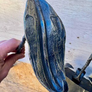 May include: A handcrafted, dark blue and gold-toned metal sculpture of a stylized leaf. The leaf-shaped art piece has a textured surface and is mounted on a metal stem. The sculpture is placed on a wooden surface.