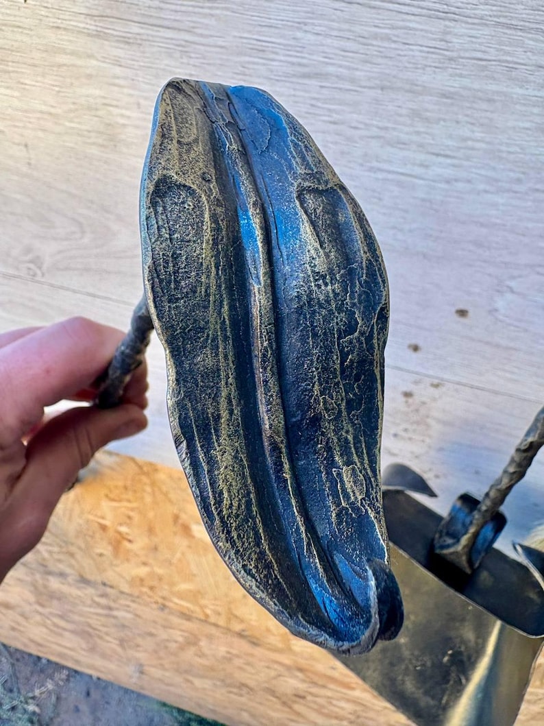 May include: A handcrafted, dark blue and gold-toned metal sculpture of a stylized leaf. The leaf-shaped art piece has a textured surface and is mounted on a metal stem. The sculpture is placed on a wooden surface.