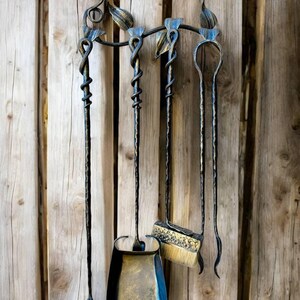 May include: A wrought iron fireplace tool set with a shovel, brush, poker, and a leaf-shaped tool. The tools are black with gold accents and are mounted on a decorative holder with leaf and swirl details. The background is a wooden wall.