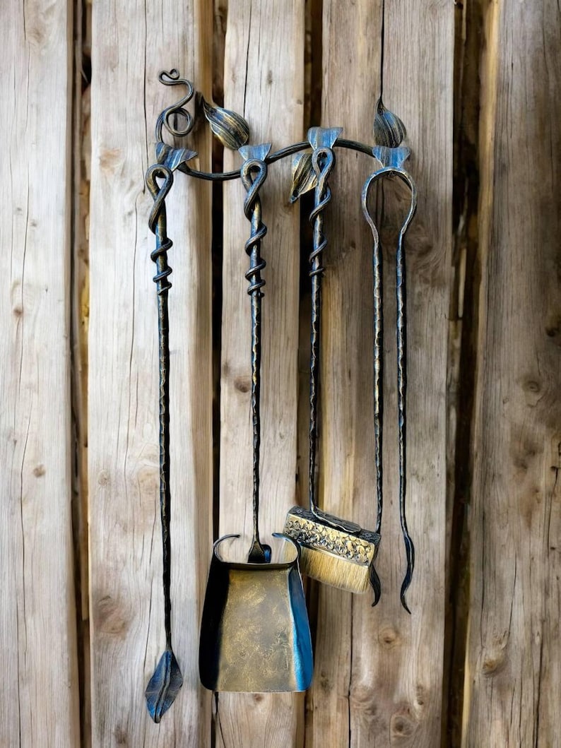 May include: A wrought iron fireplace tool set with a shovel, brush, poker, and a leaf-shaped tool. The tools are black with gold accents and are mounted on a decorative holder with leaf and swirl details. The background is a wooden wall.