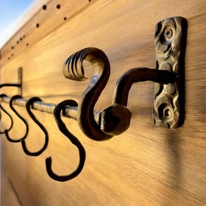 May include: Hand-forged metal wall-mounted coat rack with decorative S-hooks. The rack has a textured, dark brown finish and is mounted on a light wood surface. The design includes a curved, scroll-like detail on the end.