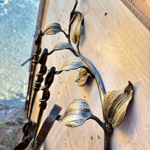 May include: Decorative metal wall art featuring a floral design with leaves and stems. The metal has a gold and bronze finish, with a textured appearance. The art is mounted on a light-colored wooden surface.