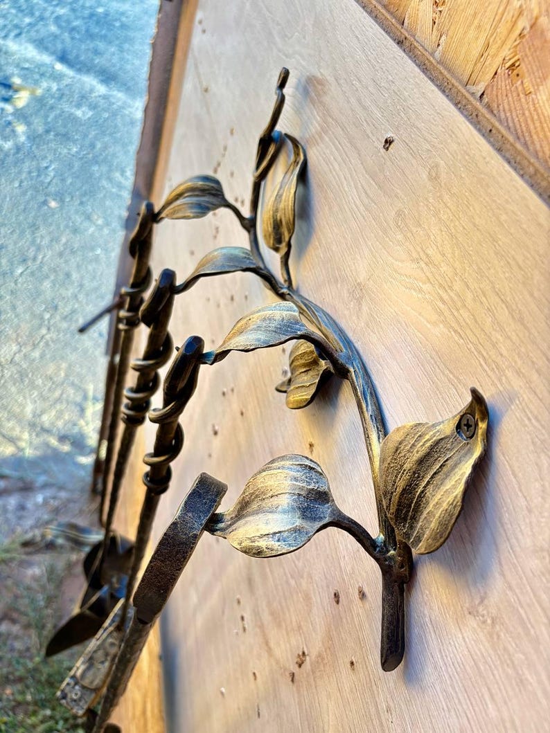 May include: Decorative metal wall art featuring a floral design with leaves and stems. The metal has a gold and bronze finish, with a textured appearance. The art is mounted on a light-colored wooden surface.
