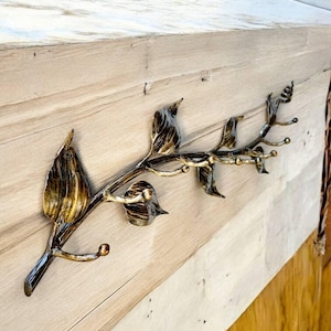 Puede incluir: Un colgador de pared decorativo de metal con forma de rama con hojas y pequeños ganchos. El metal tiene un acabado dorado y bronce. El colgador de pared está montado sobre una superficie de madera clara.