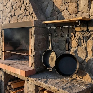 Könnte beinhalten: Outdoor-Grill aus Stein mit eingebautem Grill und Holzablage. Zwei schwarze gusseiserne Pfannen hängen an einem Schmiedeeisenregal. Brennholz ist unter dem Grill gestapelt.