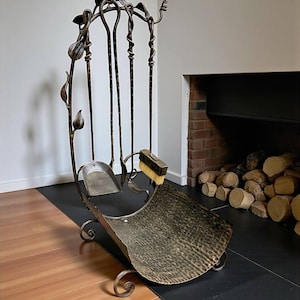 May include: A wrought iron fireplace tool set with a leaf and vine design. The set includes a shovel, poker, brush, and a log holder. The tools are arranged in a decorative stand with a textured, hammered metal finish. The set is designed for use near a fireplace.