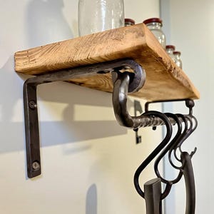 Puede incluir: Estantería rústica de madera con borde natural, soportada por soportes de metal oscuro. La estantería tiene una fila de ganchos colgantes debajo. Frascos de vidrio transparente con tapas rojas se encuentran en la estantería.