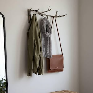 Puede incluir: Un perchero rústico montado en la pared hecho de ramas naturales, con una chaqueta verde oliva, una bufanda gris y un bolso mensajero de cuero marrón. Debajo hay un banco de madera y una cesta de mimbre.