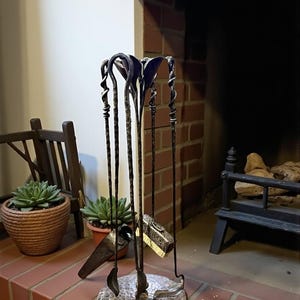Op de afbeelding: Een smeedijzeren haardgereedschapsset met een gehamerde metalen basis. De set bevat een pook, schop, borstel en tang. De gereedschappen hebben decoratieve gedraaide en gebogen handvatten. De set wordt getoond op een bakstenen haard.