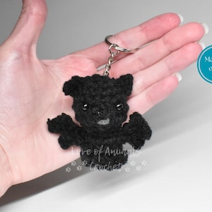 May include: A black crocheted bat keychain featuring small black plastic eyes and a tiny grey tooth. The bat is attached to a silver-colored metal split ring by a short chain. The text "Made to order" appears in a blue circle, and "Love of Animals Crochet" is printed below the bat.