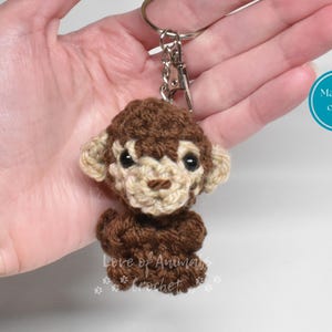 May include: Crocheted monkey keychain in shades of brown and tan. The handmade monkey has a detailed face and is attached to a silver-colored keychain. The text "Love of Animals Crochet" is visible on the bottom of the monkey.