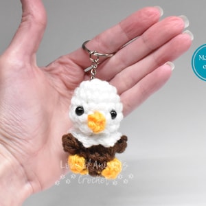 May include: A handmade crochet keychain featuring a small, adorable bald eagle. The eagle has a white head, brown body, and yellow feet and beak. The keychain is attached to a silver ring. The text "Made to order" is in a blue circle.