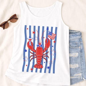Puede incluir: Una camiseta sin mangas blanca con el gráfico de una langosta roja que lleva gafas de sol en forma de estrella. La langosta sostiene una copa de martini y una bandera estadounidense sobre un fondo de rayas azules verticales, estrellas rojas y fuegos artificiales.