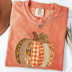 Könnte beinhalten: Ein korallenfarbenes Kurzarm-T-Shirt mit einer großen Kürbisgrafik, die aus verschiedenen Stoffflicken wie Karomustern, Vichy-Karos und Blumenmustern besteht. Das T-Shirt hat einen Rundhalsausschnitt und ein Comfort Colors-Etikett in Größe S-P-CH.