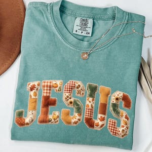 May include: A teal short-sleeve t-shirt with a crew neckline and a Comfort Colors label. The front features the word JESUS in large, block-style letters, each filled with a patchwork pattern of autumn leaves, pumpkins, and plaid in orange and brown.
