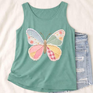 Könnte beinhalten: Ein tealfarbenes Tanktop mit einem bunten Schmetterlings-Applikation. Das Schmetterlingsdesign enthält verschiedene Muster und Farben, darunter rosa Punkte, gelbes Vichy und Blumenmuster. Das Tanktop wird mit Jeansshorts kombiniert.