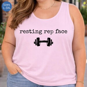 May include: A light pink tank top with the text "resting rep face" above a black dumbbell graphic. The tank top is a solid color and has a scoop neck. Sizes L-3XL are available.
