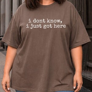 Puede incluir: Una camiseta marr&oacute;n de manga corta con el texto blanco "i dont know, i just got here" impreso en una fuente estilo m&aacute;quina de escribir en el pecho. La prenda tiene un cuello redondo y un corte relajado, ofreciendo un aspecto sencillo.