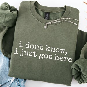 Puede incluir: Una sudadera de cuello redondo en color verde oliva con el texto blanco "i dont know, i just got here" impreso en la parte delantera. La prenda tiene un cuello acanalado y mangas largas, y un collar de cadena plateada sobre el escote.