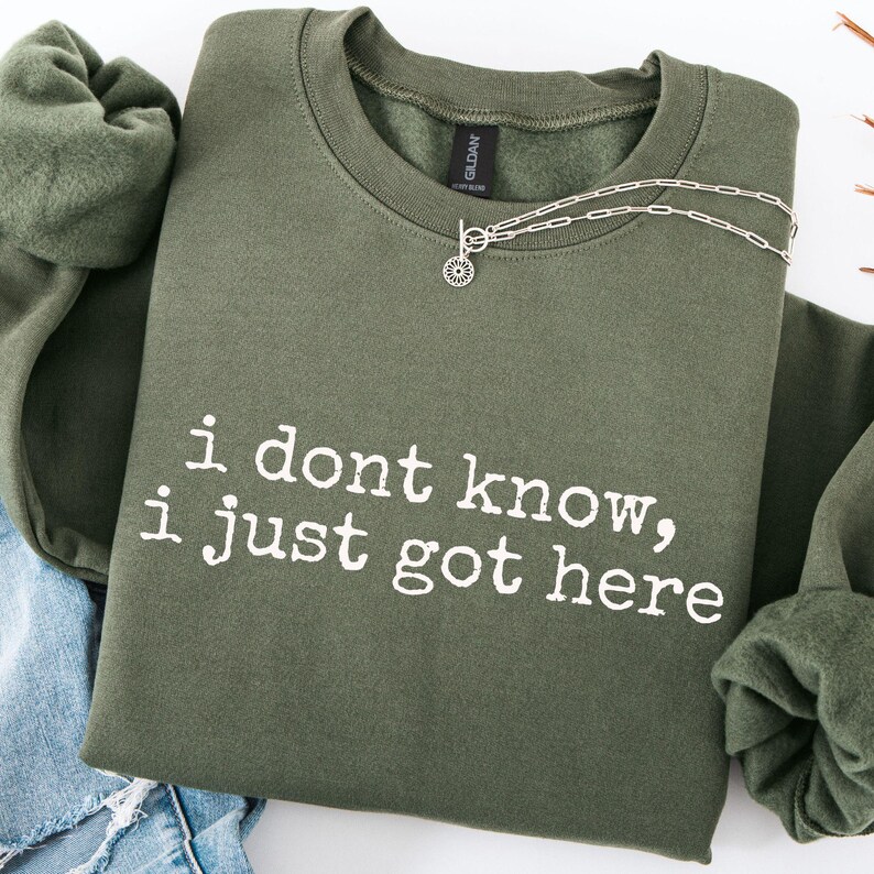 Puede incluir: Una sudadera de cuello redondo en color verde oliva con el texto blanco "i dont know, i just got here" impreso en la parte delantera. La prenda tiene un cuello acanalado y mangas largas, y un collar de cadena plateada sobre el escote.