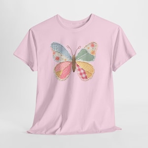 May include: A light pink short-sleeve t-shirt featuring a central graphic of a butterfly. The butterfly wings are composed of various fabric patches in pastel colors, including blue gingham, floral prints, pink polka dots, and red checkered patterns.